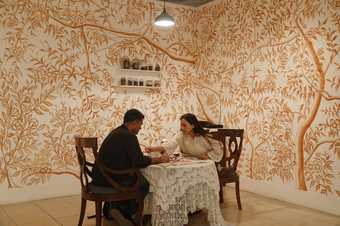 two people sit at a table covered in a lace cloth in a room with floral wallpaper