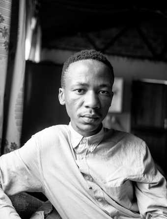 A black and white photograph of the artist Lindokuhle Sobekwa showing his head and torso, wearing a light-coloured button-up shirt