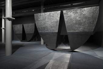 A photograph of of Tate Liverpool's ground floor gallery which features Torkwase Dyson's huge sculptures. These are large semi-circular pieces which appear to balance on the curved edge. They are black with a triangular area of the semi-circle cut out.