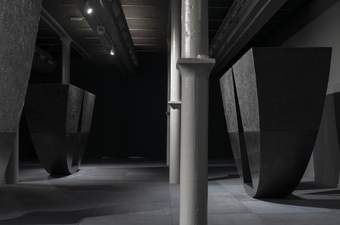 A wide photograph of Tate Liverpool's ground floor gallery which features Torkwase Dyson's huge sculptures. These are large semi-circular pieces which appear to balance on the curved edge. They are black with a triangular area of the semi-circle cut out.