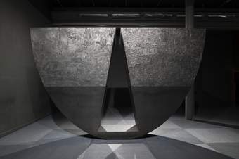 A photograph of one of Torkwase Dyson's huge sculptures. It is a large semi-circular shape which appears to balance on the curved edge. There is a triangular area of the semi-circle cut out and the sculpture is black.