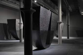 A wide photograph of Tate Liverpool's ground floor gallery which features Torkwase Dyson's huge sculptures. These are large semi-circular pieces which appear to balance on the curved edge. They are black with a triangular area of the semi-circle cut out.