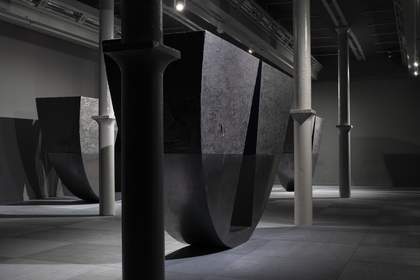 A wide photograph of Tate Liverpool's ground floor gallery which features Torkwase Dyson's huge sculptures. These are large semi-circular pieces which appear to balance on the curved edge. They are black with a triangular area of the semi-circle cut out.