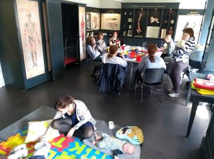 People and babies are seated in a room by a table and on the floor.