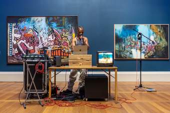 A person with a microphone, laptop and sound system performing in front of abstract paintings