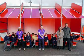 A photograph of the Mobile Museum art truck with a group of school children sat on the benches in front with guides of the exhibition, Tate Liverpool Director Helen Legg and entrepreneur and philanthropist Frederic Jousset