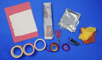 Materials laid out on a table, with a blue background. Including: Scissors, glue, card, ribbon, shiny packaging and fruit netting