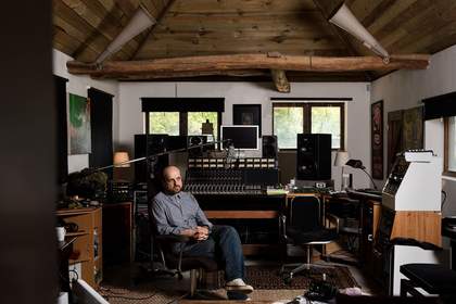 The artist sits in a chair in a music studio filled with sound equipment to advertise the event.
