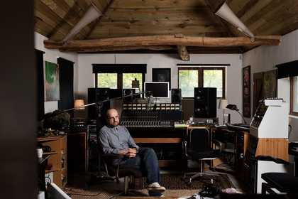 The artist sits in a chair in a music studio filled with sound equipment to advertise the event.