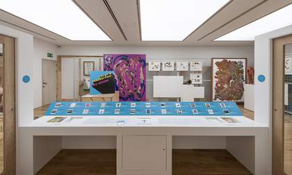 A museum gallery containing a white display table with images and interpretation text in the foreground and a metal mesh pull out screen supporting hanging framed artworks in the background.