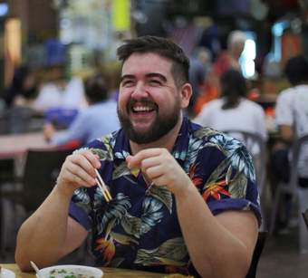 A man in a patterned top, with brown hair and beard, holding a pair of chopsticks.