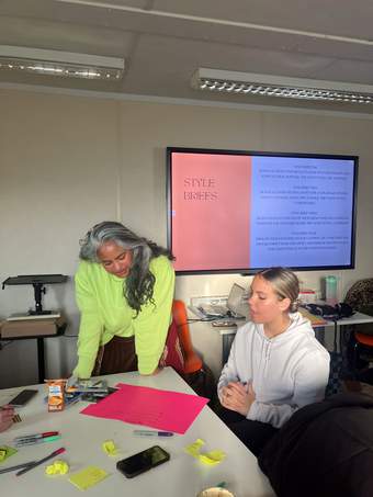 A photograph of a young person talking with stylist Mithila Ramagavigan, both looking at a pink piece of paper on the table.