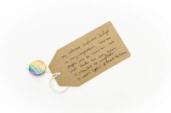 Photograph of a badge which says ‘LGBTI intersex inclusive’, with a brown paper label attached, with handwritten text