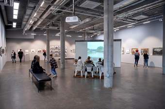 A landscape overview image of Northern Gallery Contemporary Arts' gallery space, with a crowd seated in the centre, watching a projector screen, two people seated on a bench and multiple people in the background viewing the framed art on the walls