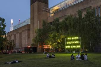 people sit on the lawn outside Tate Modern by a neon green sign that says 'we must cultivate our garden'