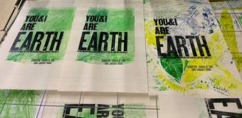Screenprints on a drying rack. The prints have green, leafy backgrounds, with text saying ‘You and I are Earth’