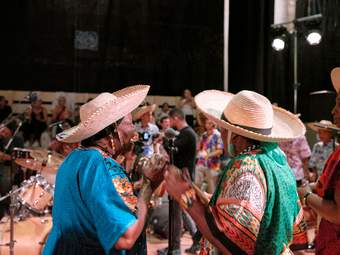 Two womnen in colorful attire and hats perform on stage and look at eachother