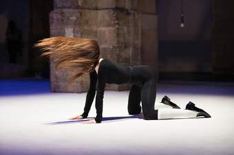 A dancer posing on all fours on a white stage, wearing a black top and leggings with white socks, swinging their long hair