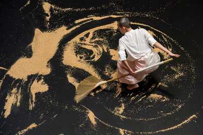 An image taken from above of a dancer dressed in a white shirt and pink/white sarong. The dancer is crouching on a black floor scattered with brown rice which has been swept into labyrinthine swirls. The dancer dynamically holds a broom behind their back