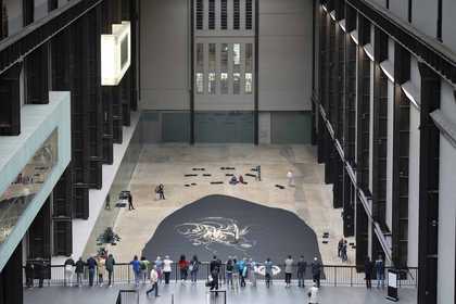 wide view of a large industrial gallery space with audiences watching performers sweeping rice on a dark floor