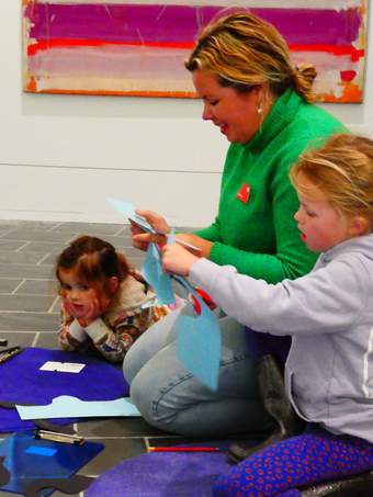 Woman in green jumper with two young girls either side of her. They sit in front of a colourful painting, cutting card to make art together.