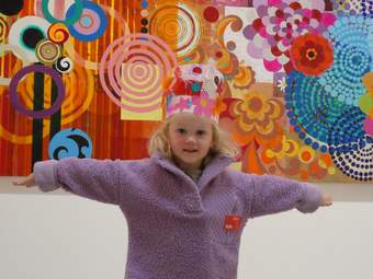 Young girl with arms outstretched, wearing colourful headdress, in front of bright, patterned artwork