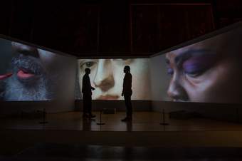 2 people silhouetted against a 3-channel film installation, featuring from left to right, close up shots of a Black bearded person applying lipstick, a painted portrait of a white person, and an a Black person with purple eyeshadow