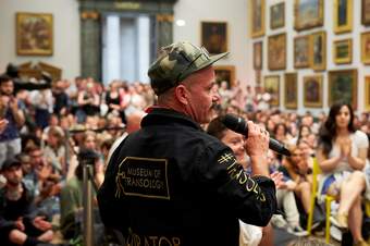Photograph of E-J Scott, wearing a black boiler suit with ‘Museum of Transology’ embroidered across the back, speaking into a microphone, with a room full of people in the background
