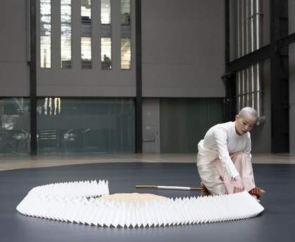 performer moving a paper construction encircling a mound of rice on a dark floor