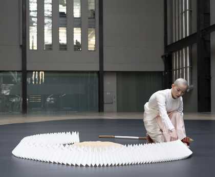 performer moving a paper construction encircling a mound of rice on a dark floor