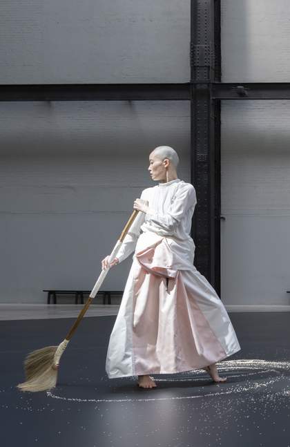 performer holding broom standing on a dark surface scattered with rice