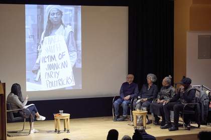 A panel event on a stage with a person interviewing four panellists who are all seated. A large screen projects a photograph of a woman holding a placard that reads 'Michael Smith: Victim of Jamaican Party Politricks!'