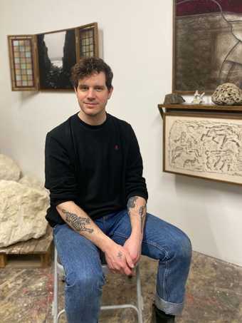 Man sat on a chair, wearing jeans and jumper, artworks on the walls behind him.