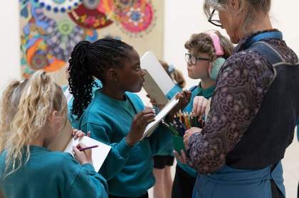 A group of children in school uniforms holding sketchbooks. An adult hands out coloured pencils.