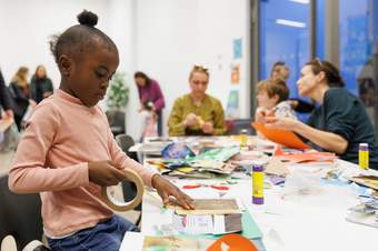 Girl sticks paper down at a table with other families in an art workshop at Tate