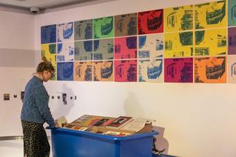 A photograph of a visitor wearing a blue jumper looking at a display of books on a blue trolley. There is a series of colourful prints displayed on the wall above.