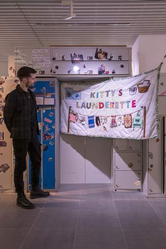 A photograph of a visitor standing in RESOLVE Collective's exhibition looking at a material banner with the words 'Kitty's Laundrette' embroidered on it.