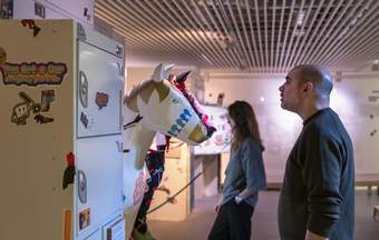 A photograph of a visitor looking at two sculptures of horse's heads. There is a locker on the left hand side covered in stickers and there is a visitor in the background wearing headphones.