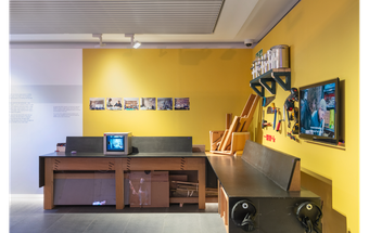 A photograph of RESOLVE Collective's display at Tate Liverpool + RIBA North. It features a work bench with different tools and a tv monitor. The walls behind are painted yellow.