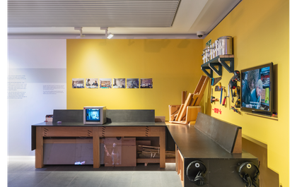 A photograph of RESOLVE Collective's display at Tate Liverpool + RIBA North. It features a work bench with different tools and a tv monitor. The walls behind are painted yellow.
