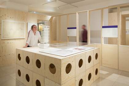 A photograph of visitors in an exhibition space surrounded by wooden structures and architectural models