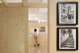 A photograph looking through a gap between two wooden walls at a visitor who is looking at an artwork on the back wall.