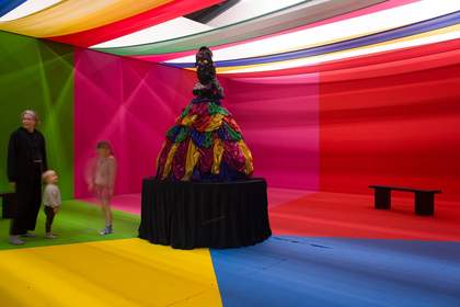 Image shows at the centre, a large sculpture of a black female figure in a layered, colourful skirt standing on a black pedestal. It is situated in a room with brightly coloured hanging fabric on the ceiling and walls, with the floor painted in the same c