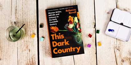 A book against a wooden background with a glass of water, paints and a palette.
