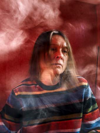Photograph of Sarah Lucas in motion surrounded by swirls of smoke on a red background