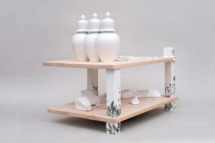 Three white porcelain vessels on a table like structure with three wooden legs painted white with fern patterns and a lower lever with lumps of white porcelain