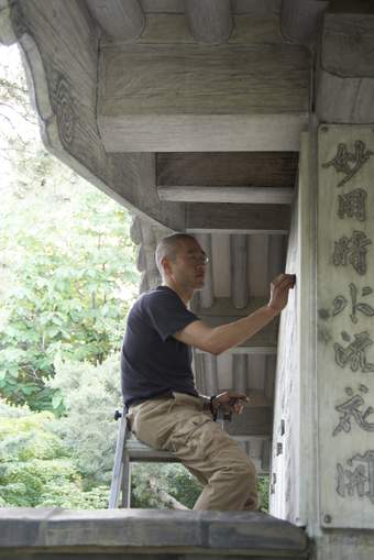 A man sitting on the top of a ladder outside a house, raising his hand to draw something on the wall of the building.