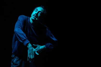 A man moving in a dynamic motion, pressing his arms outwards, with his eyes closed. He is lit by a soft glowing blue light. He is wearing black clothes and has a microphone attached round his head.