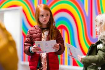 Child in red coat standing centre of frame holding Art Explorer card looking around gallery. Smaller child right of frame holding Art Explorer card. Background is brightly coloured wall mural.