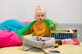 Small child in yellow jumper playing with green scarf, sitting amongst colourful bean bags and treasure baskets.